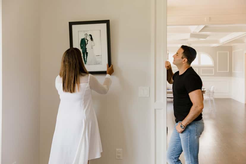 Two people hanging a framed black and white portrait on a hallway wall at eye level