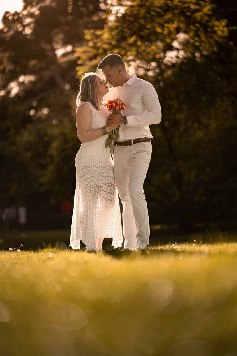 A couple embracing face to face outdoors at sunset in a park, the man in a white shirt holding a bouquet of orange tulips between them