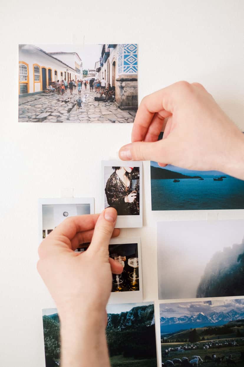 Two hands pinning a small photo print onto a wall that already holds several other photo prints, building a gallery wall