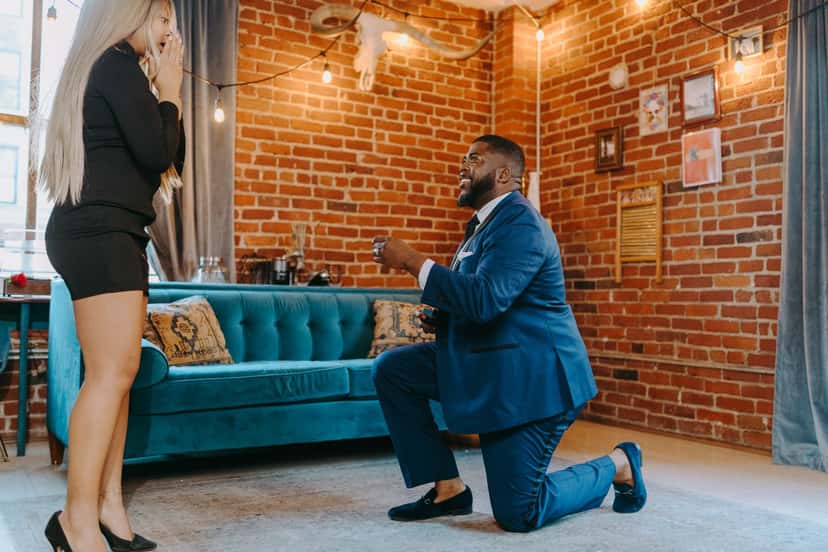 A man in a blue suit kneeling on one knee to propose, presenting a ring to a woman in a black dress who covers her mouth in surprise, in a living room with an exposed brick wall and a teal sofa